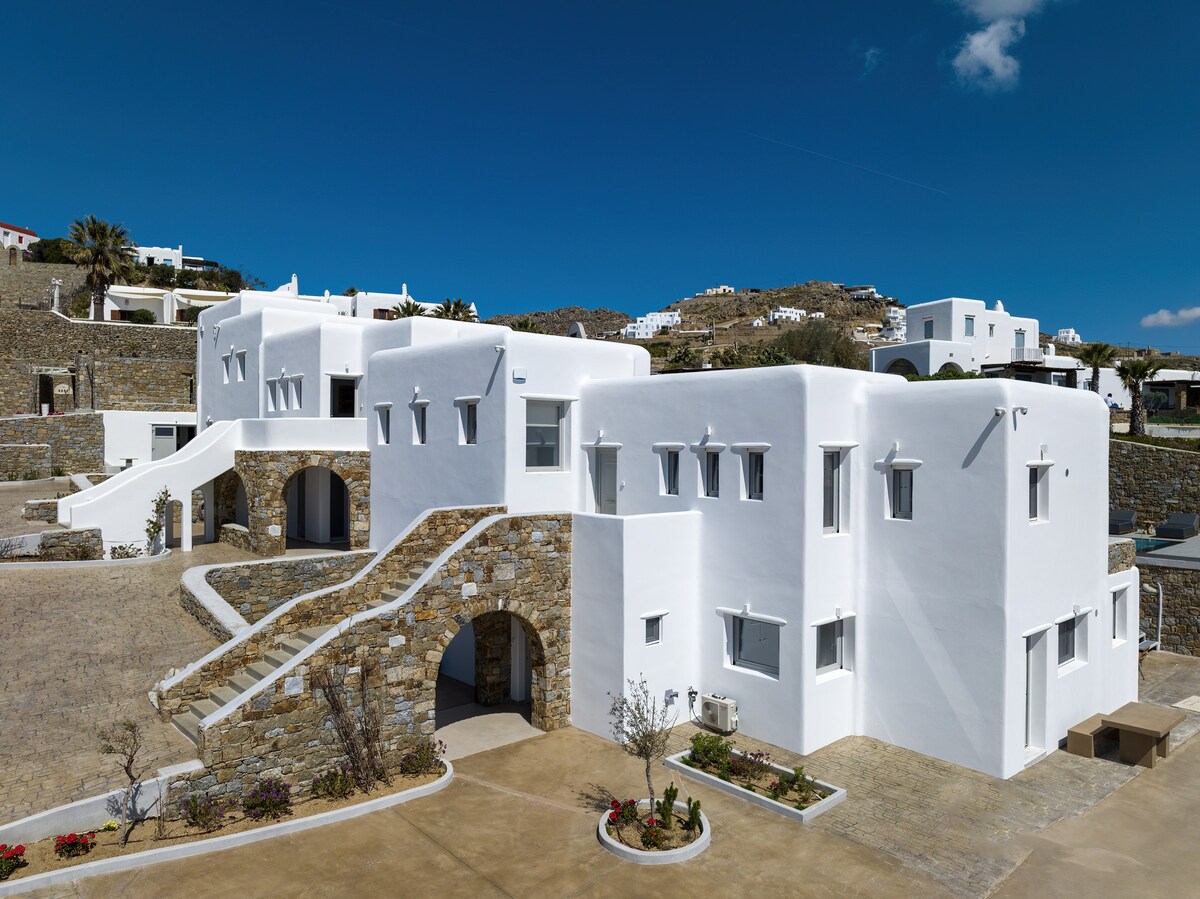 A modern villa complex features a blend of whitewashed buildings and natural stone elements. The exterior showcases clean lines and multiple arches, with landscaped areas enhancing the entrance. Clear blue skies provide a bright backdrop, highlighting the serene and minimalist architectural design.