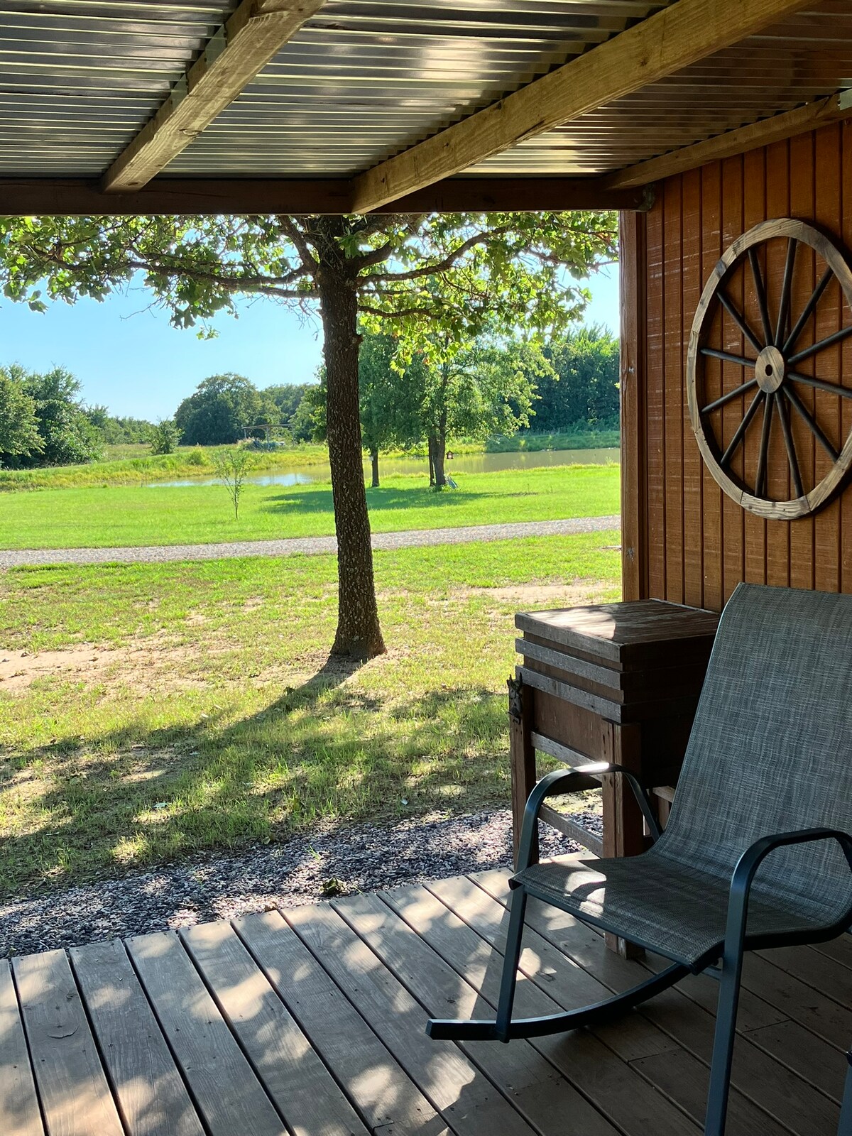 A shaded porch area is shown, featuring a comfortable chair and a small table. In the background, green fields and trees are visible, with a winding path leading to a pond that reflects the clear sky. A rustic decoration resembling a wagon wheel adorns the wall.