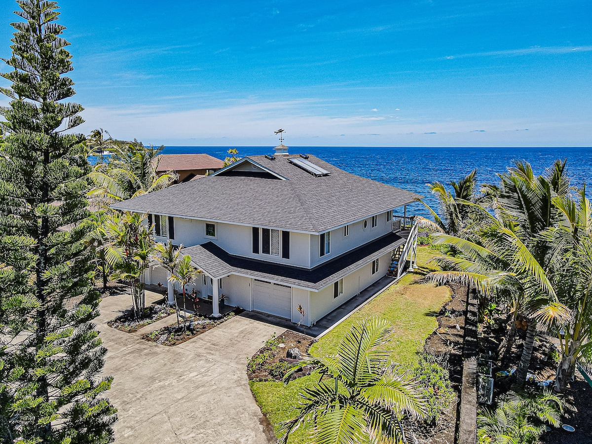 The exterior of the rental is shown, featuring a two-story structure with a sloped roof. Palm trees and lush landscaping surround the property, which is adjacent to the ocean. A clear blue sky complements the tranquil ocean view visible in the background.