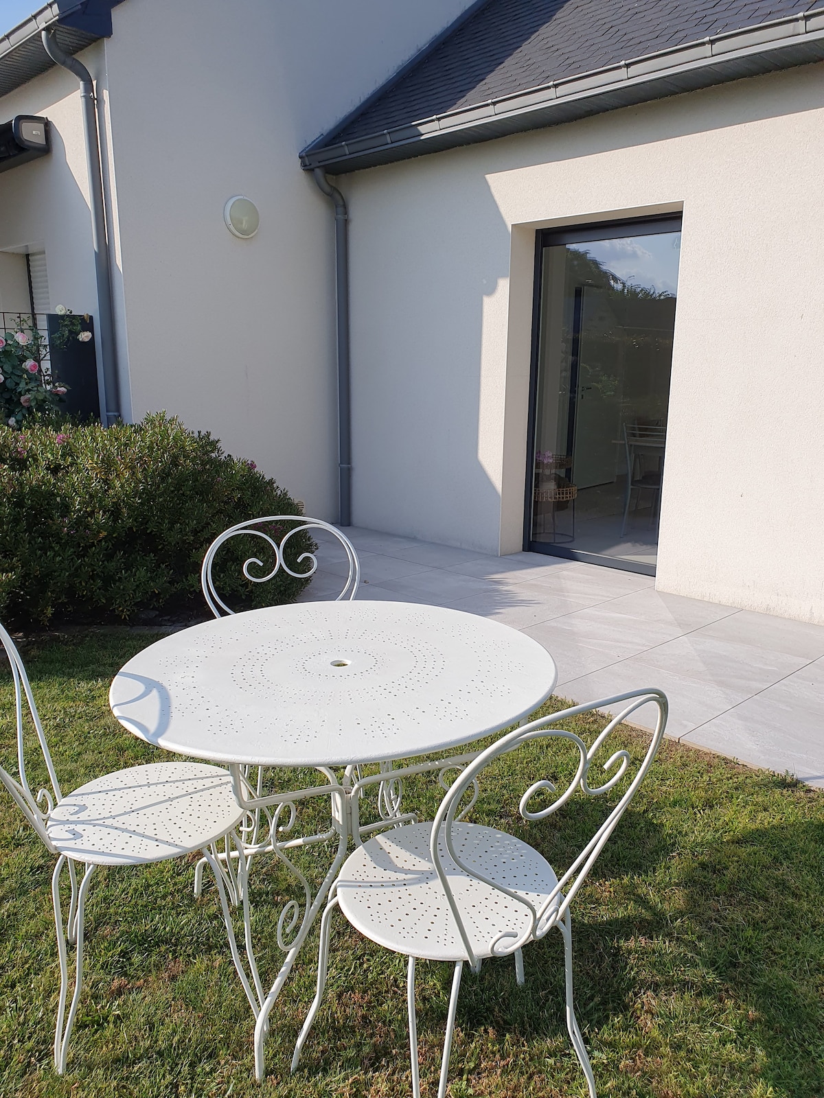 An outdoor seating area is presented with a round white table surrounded by three matching chairs. The setting is situated on a grassy patch near a stone patio, leading to sliding glass doors that open to the interior. Lush greenery enhances the tranquil atmosphere.