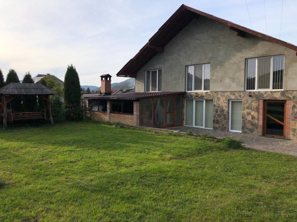 A spacious house is depicted, showcasing a combination of stone and stucco exteriors. Large windows provide ample natural light, and a covered outdoor seating area with a wooden structure is visible in the garden, which features green grass and a backdrop of gentle hills.