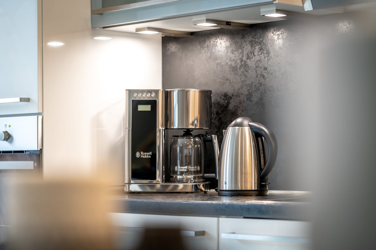 A coffee maker and kettle are placed on a kitchen countertop, with a sleek, modern design. The coffee maker features a glass carafe and digital controls, while the kettle has a metallic finish. Soft lighting enhances the overall appearance of the kitchen space.