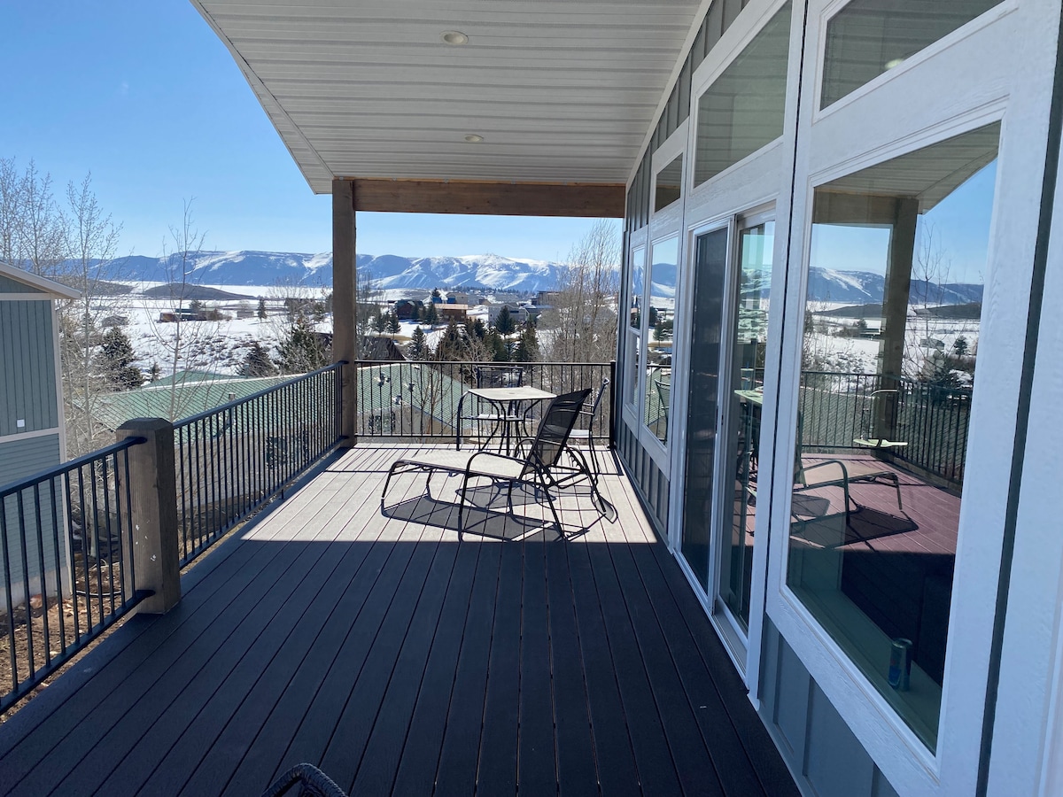 A large deck with a spacious seating area is showcased, featuring a round table and several chairs. Expansive views of snow-covered mountains and a clear sky are visible in the background, while sunlight casts shadows across the deck's surface.