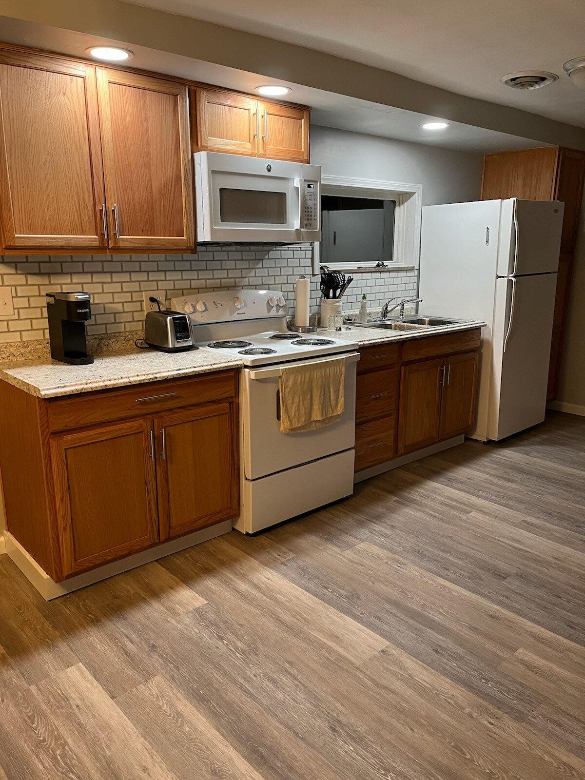 The kitchen showcases wooden cabinetry above and below a granite countertop. Appliances include a microwave, coffee maker, and refrigerator. A stove with an oven is positioned beside the sink. Wood-like flooring complements the modern design, while bright overhead lighting enhances the workspace.