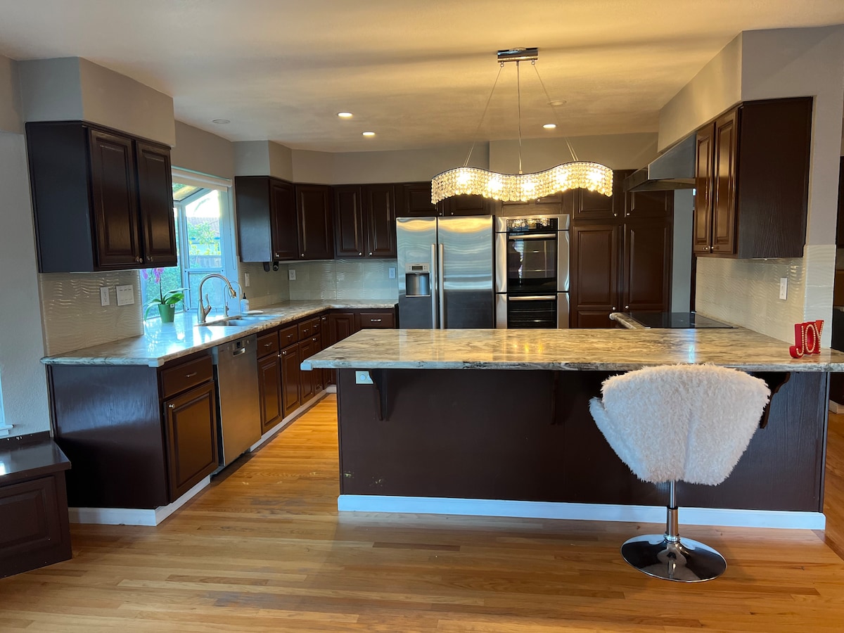 A modern kitchen is featured with dark wood cabinets and light countertops. Stainless steel appliances, including a refrigerator and oven, are prominently displayed. A unique chandelier hangs above the central island, accompanied by a plush bar stool, while hardwood flooring extends throughout the space.