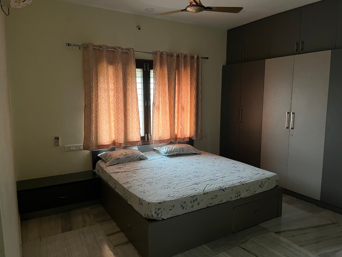 A serene bedroom is furnished with a spacious bed featuring a light floral-patterned mattress. Soft, warm light filters through the sheer curtains, while a large wardrobe and a bedside unit offer practical storage solutions.