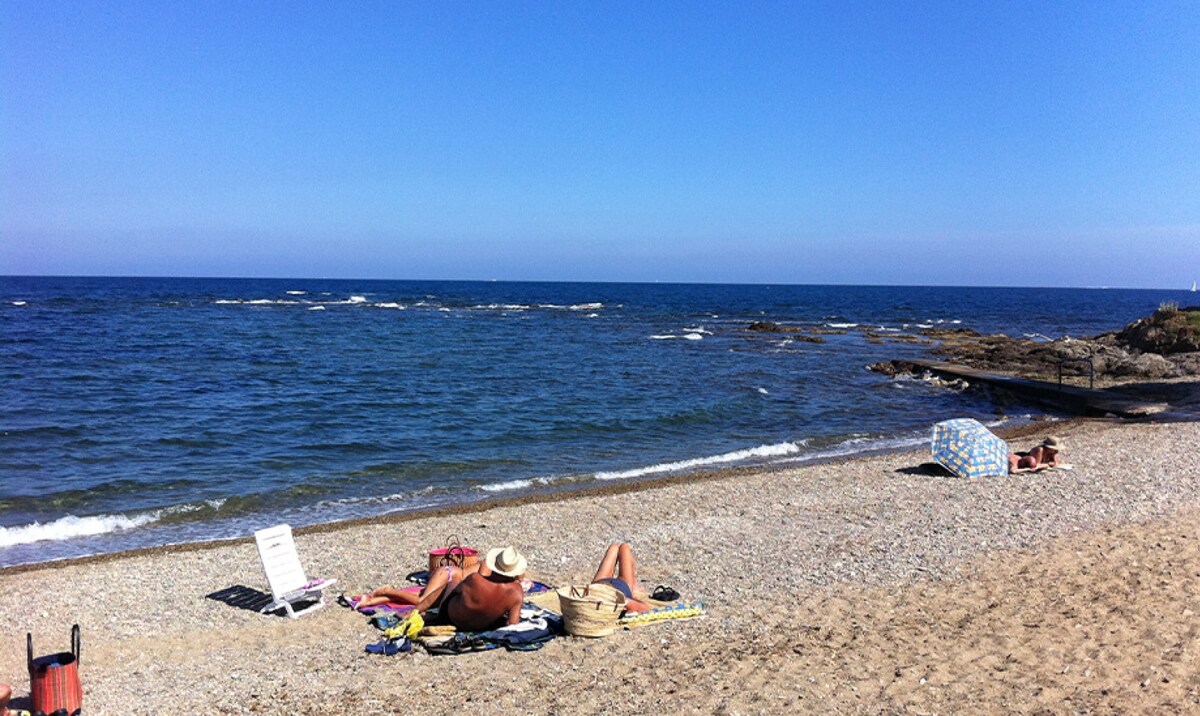 A sandy beach is depicted under a clear blue sky, with gentle waves lapping at the shore. Sunbathers relax on towels spread along the shoreline, while rocky formations and a distant beach chair are visible. The scene conveys a calm coastal environment.
