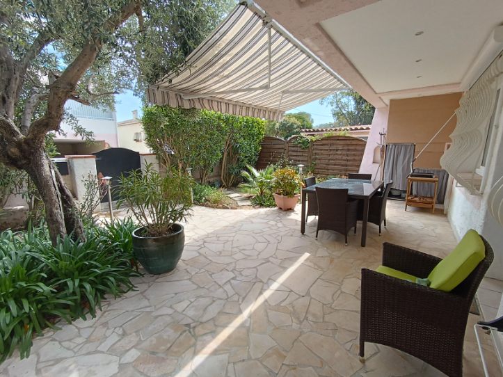 A private garden area is shown, featuring a spacious stone patio and outdoor dining table surrounded by greenery. A large awning provides shade, while potted plants and a comfortable chair enhance the tranquil setting.
