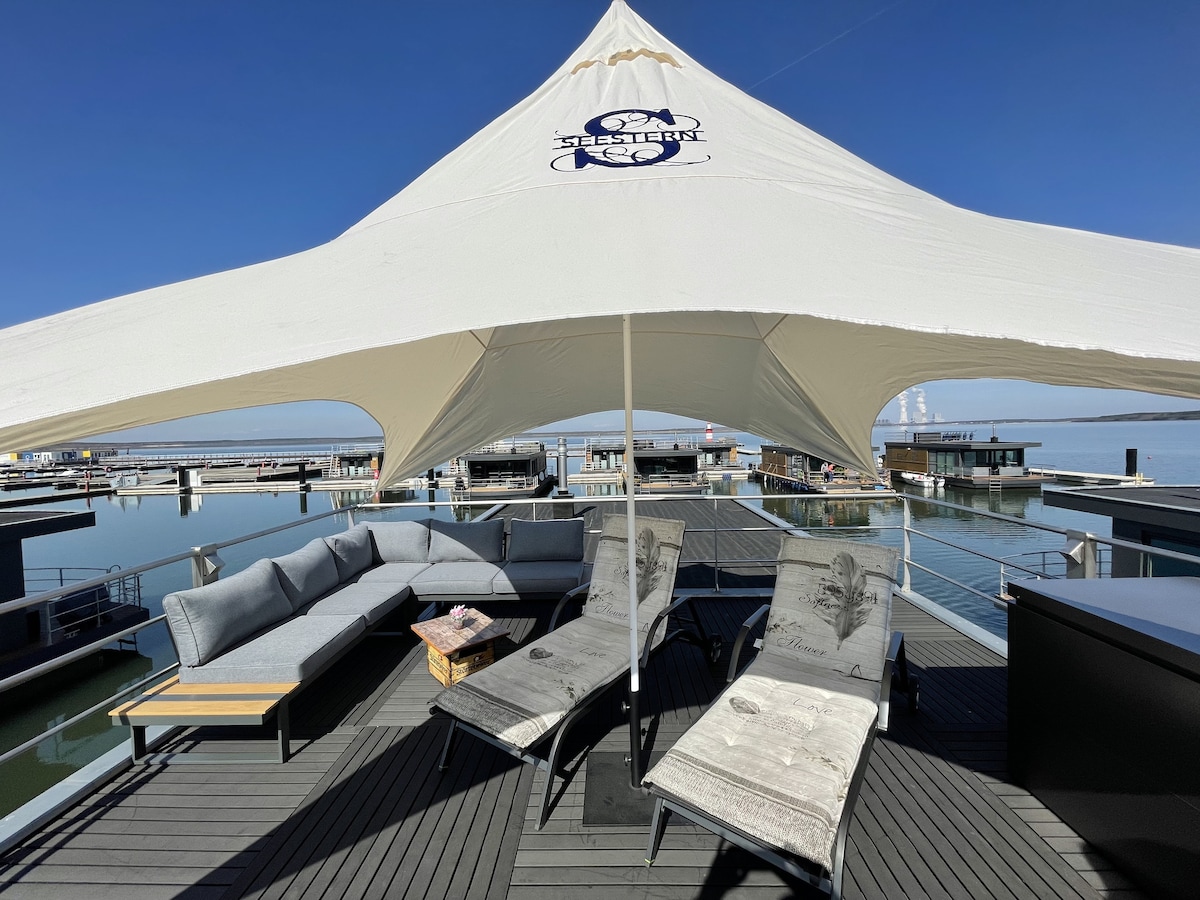 A spacious outdoor area is covered by a large canopy, providing shade over comfortable seating options. Soft seating and lounge chairs are arranged for relaxation, while a low table offers a place for drinks or snacks. The surrounding water reflects the bright blue sky.