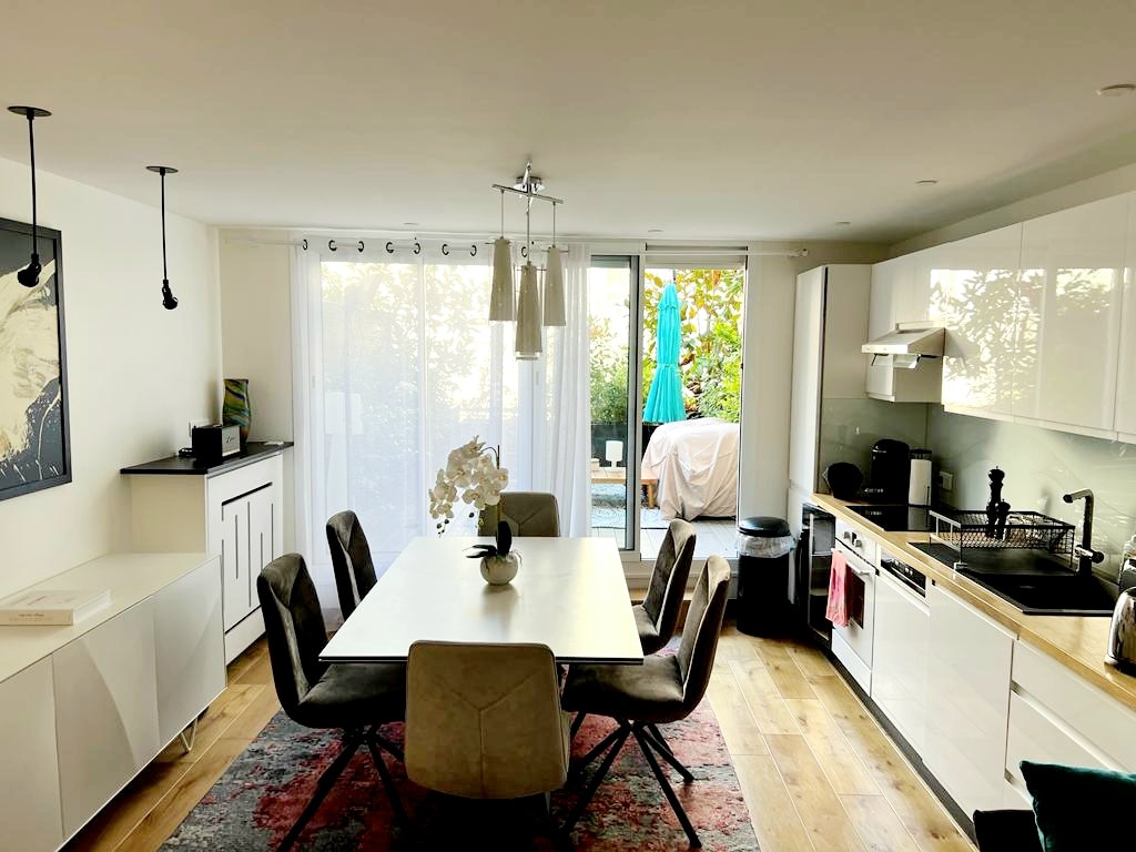 A dining area features a large table surrounded by six upholstered chairs. Natural light filters through sheer curtains, illuminating the space. In the background, a private terrace is visible, adorned with greenery and a blue lounge chair.