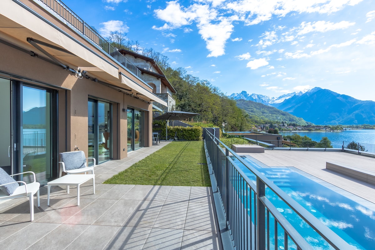 An expansive outdoor terrace overlooks the scenic Lake Como, framed by majestic mountains. Sunlight illuminates the area, which features comfortable seating and a well-maintained lawn. A shared swimming pool is visible, inviting guests to enjoy the tranquil surroundings.