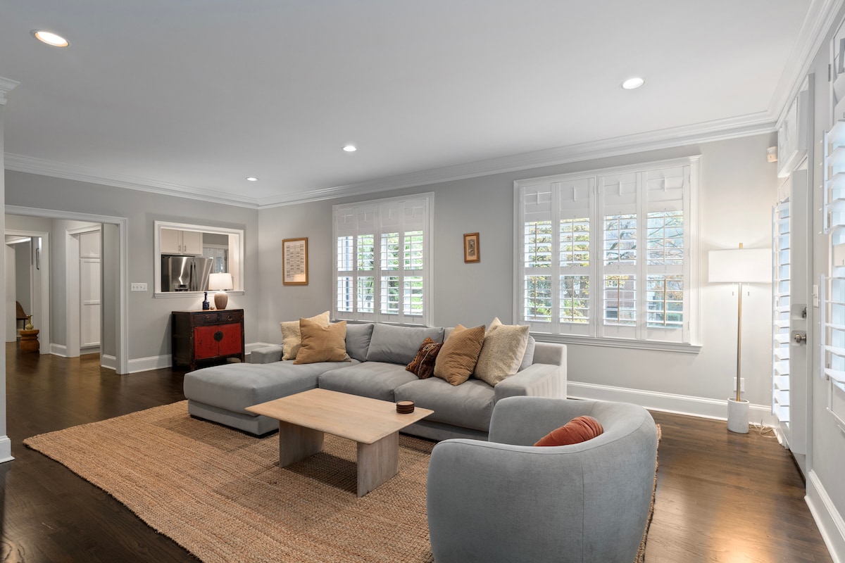 A spacious living room features a light gray sectional sofa complemented by warm-toned throw pillows. A neutral-toned area rug covers the hardwood floor. Natural light filters through the plantation-shuttered windows, illuminating a modern coffee table at the center and a wall-mounted television nearby.