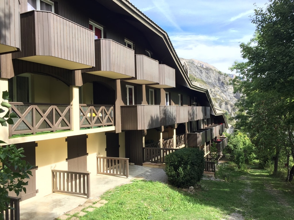 A series of chalet-style buildings showcases multiple balconies with wooden railings. The natural surroundings feature trees and shrubbery, while a pathway leads through the landscape, providing access to the entryways below. The mountains rise in the background, completing the scenic view.