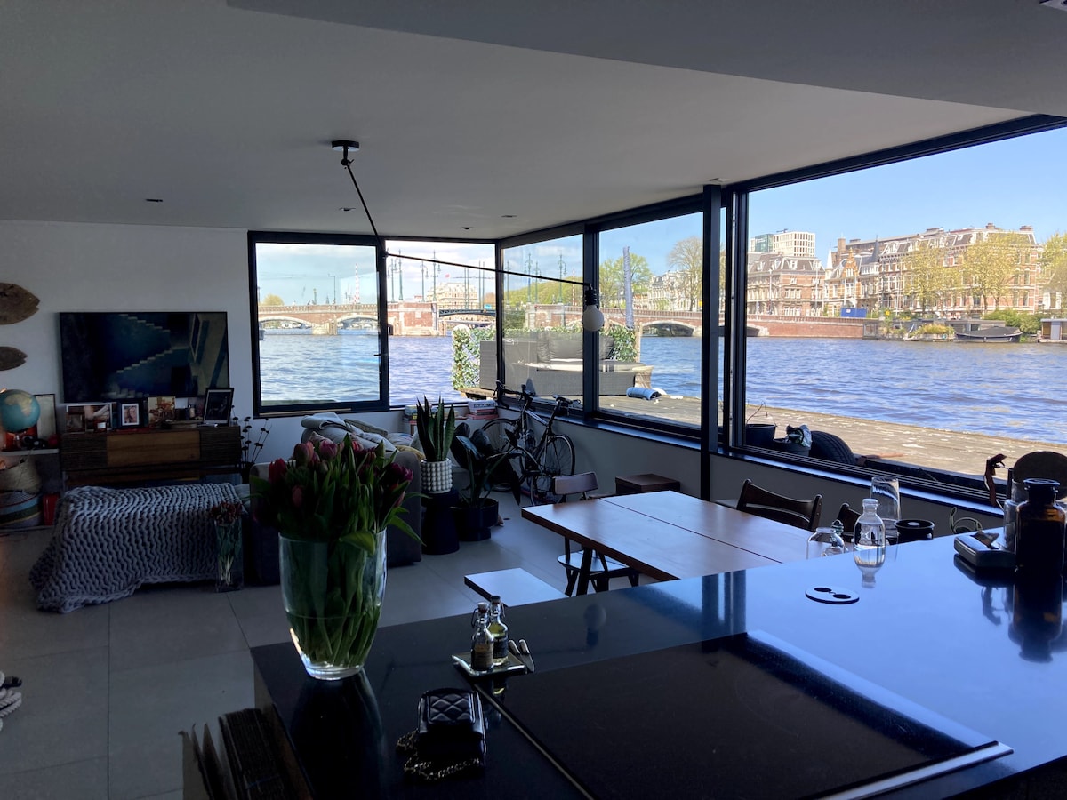 A modern living space features large glass walls that provide a view of the Amstel River. A dining table is positioned in the center, surrounded by chairs, with a vase of tulips visible. The area includes a comfortable seating arrangement and hints of outdoor scenery through the windows.