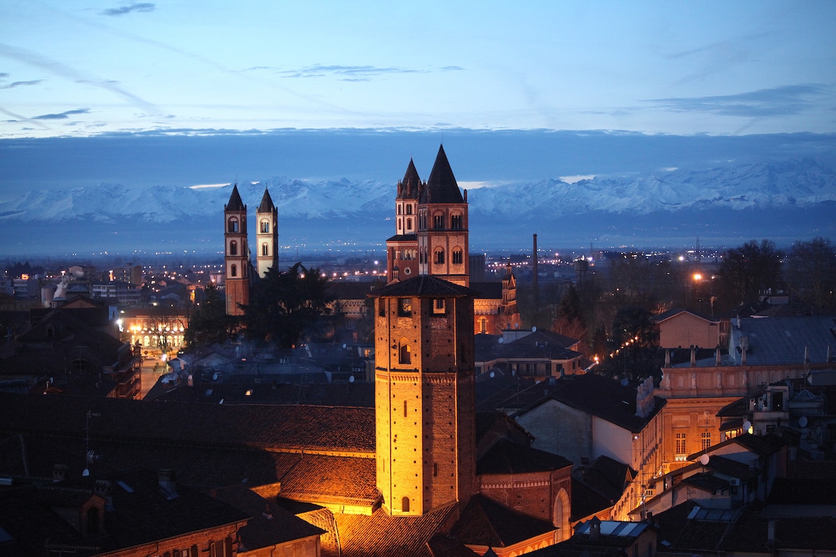 The city skyline features historic towers illuminated against a dusky sky, with distant mountains visible on the horizon. A blend of architecture showcases the charm of the area, while street lights begin to twinkle, enhancing the evening ambiance.