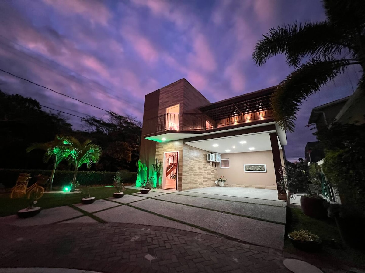 The exterior of the house is captured at twilight, showcasing a modern architectural design with warm lighting illuminating the entrance. Lush greenery and palm trees frame the property, while a paved driveway leads to the spacious entrance, enhancing the tranquil evening atmosphere.