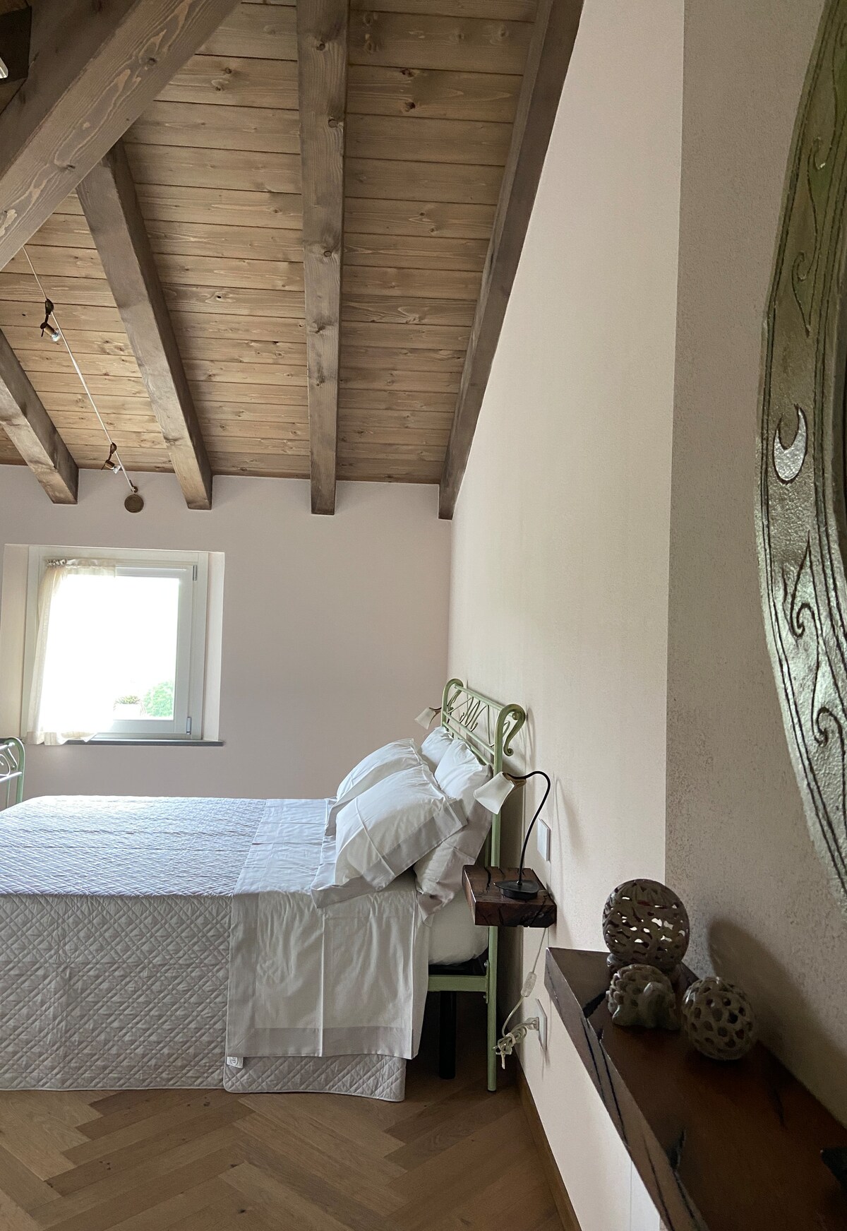 A serene bedroom featuring a spacious bed with crisp white linens and soft pillows. Natural light filters through a nearby window, illuminating the wooden ceiling beams. Decorative items, including unique sculptures, are placed on a shelf, adding a touch of personality to the space.