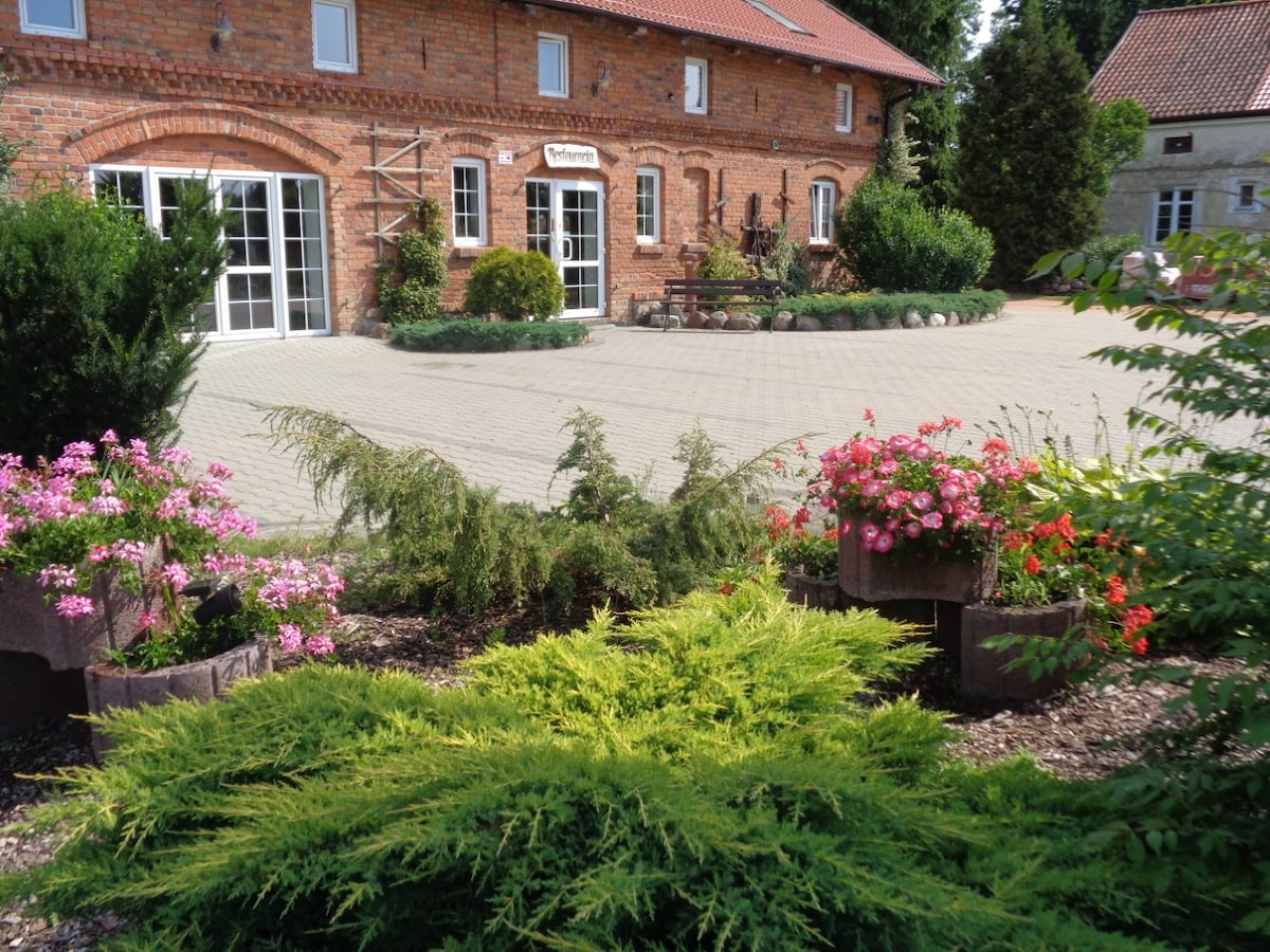A charming rustic building featuring a red brick façade and large glass doors. The landscaped area includes a variety of colorful flowers and lush greenery, with pathways of brick pavers leading to the entrance, creating an inviting outdoor atmosphere.