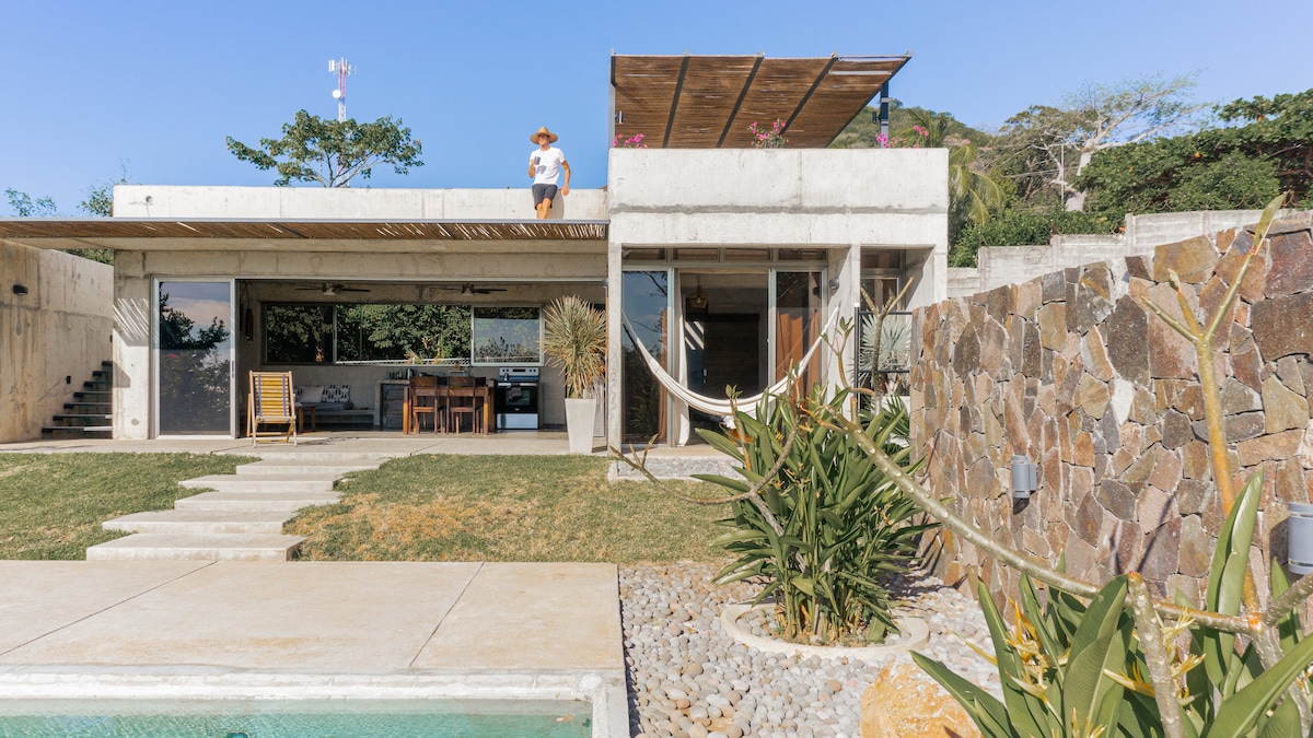 A modern house with a minimalist design is seen amidst greenery. The exterior features large glass doors that invite natural light. A rustic pool area with stone detailing is complemented by tropical plants. A seating area leads to a rooftop space framed by a wooden pergola.