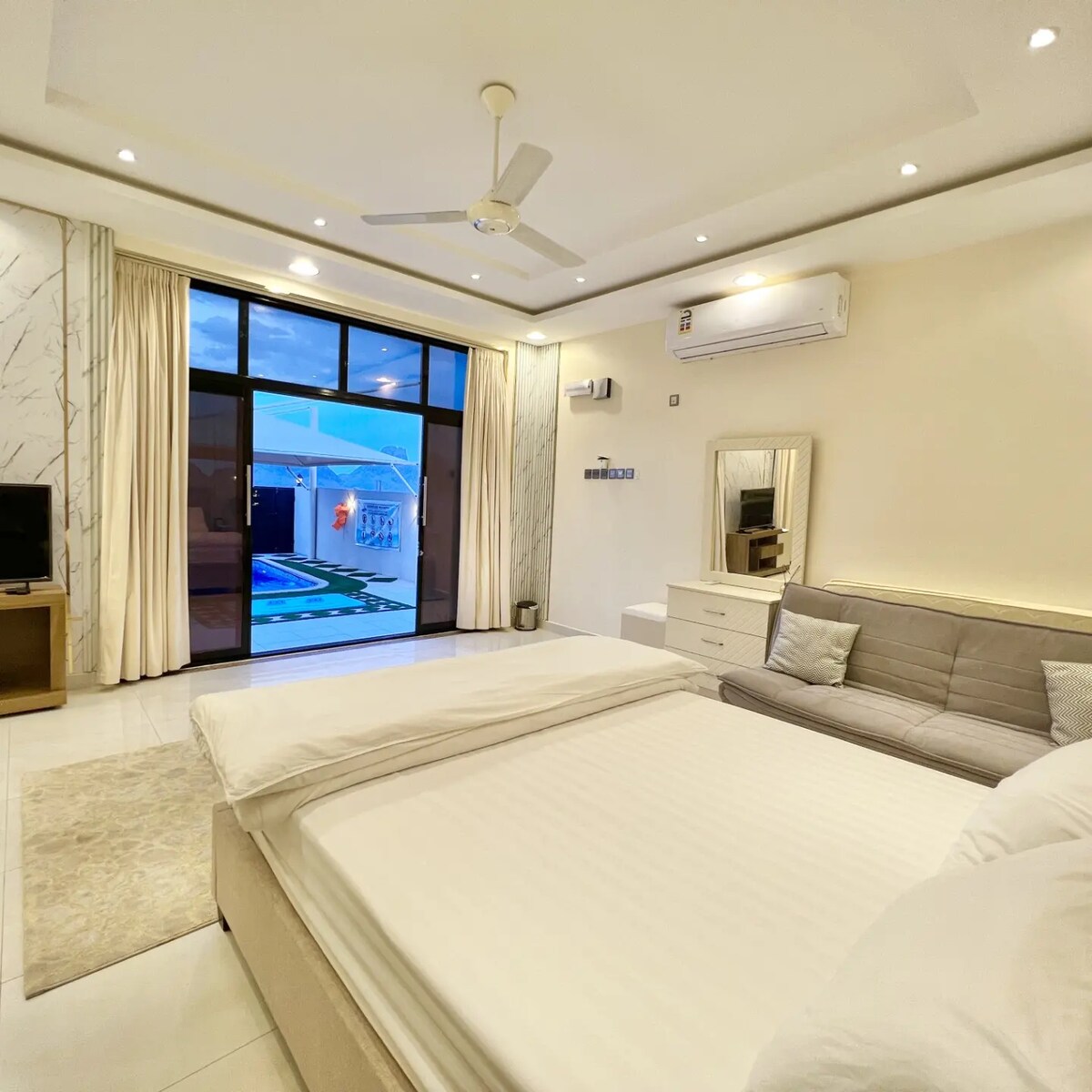 A spacious bedroom contains a plush bed and a modern sofa. Natural light floods the area through large glass doors leading to an outdoor space with a swimming pool. A television and a stylish dresser are also present, enhancing the room's functionality.