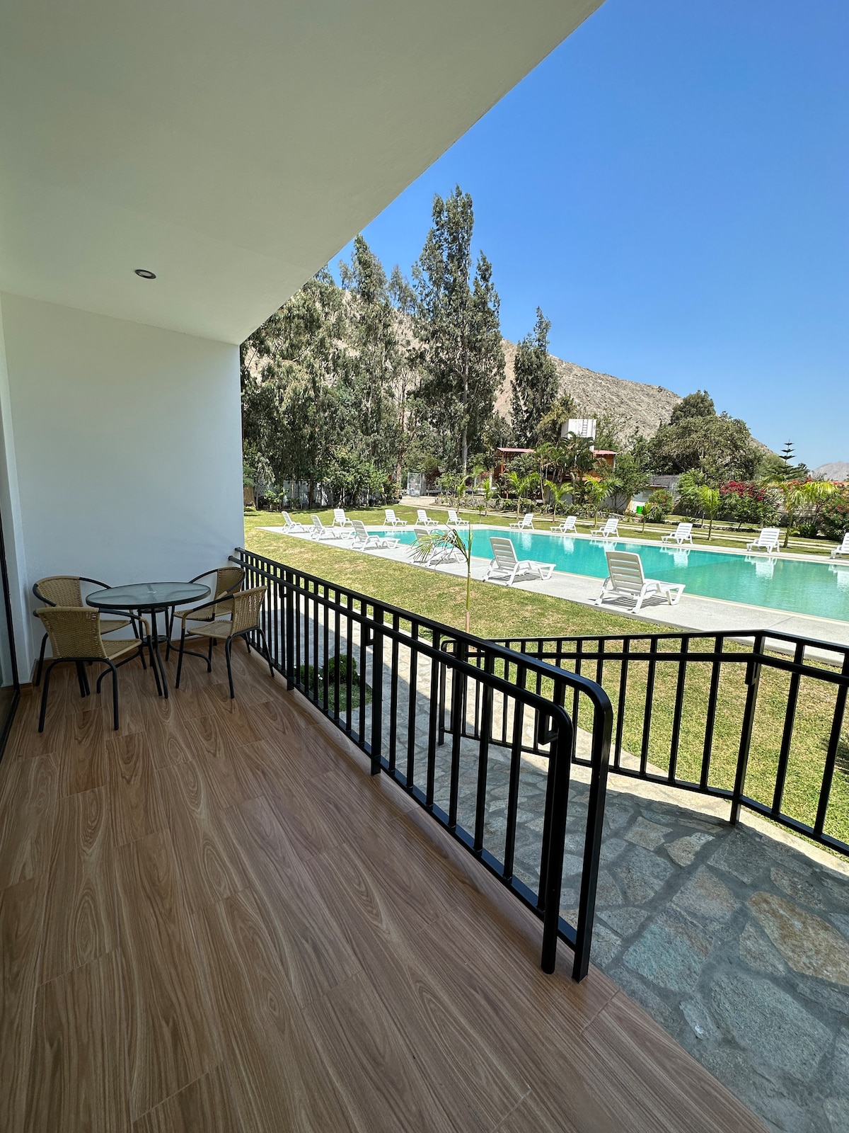 A covered balcony with a metal railing presents a view of a calm pool area surrounded by green lawns. Two wicker chairs and a small table offer a space to relax while overlooking the well-maintained grounds and mountains in the background under a clear blue sky.