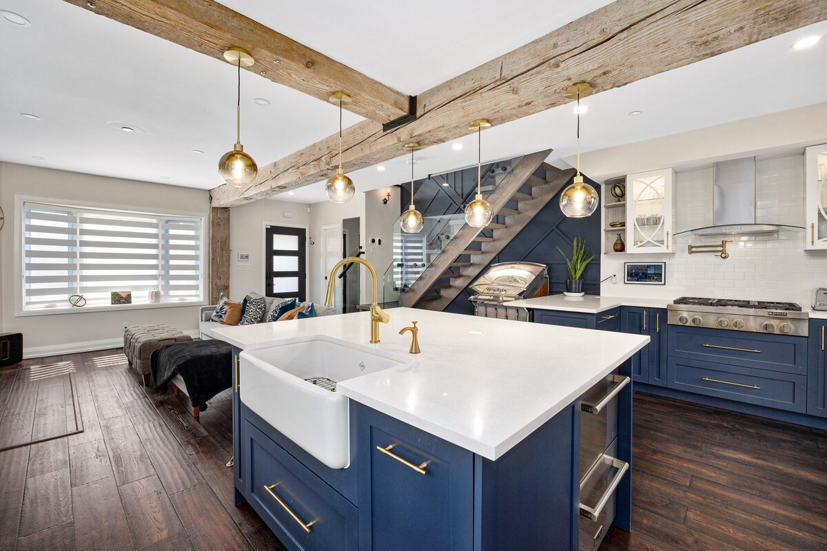 A spacious kitchen area features a large island with a farmhouse sink and gold pendant lighting. The blue cabinetry contrasts with white countertops, while a staircase with a modern design is visible in the background. Natural light pours in through the window coverings.