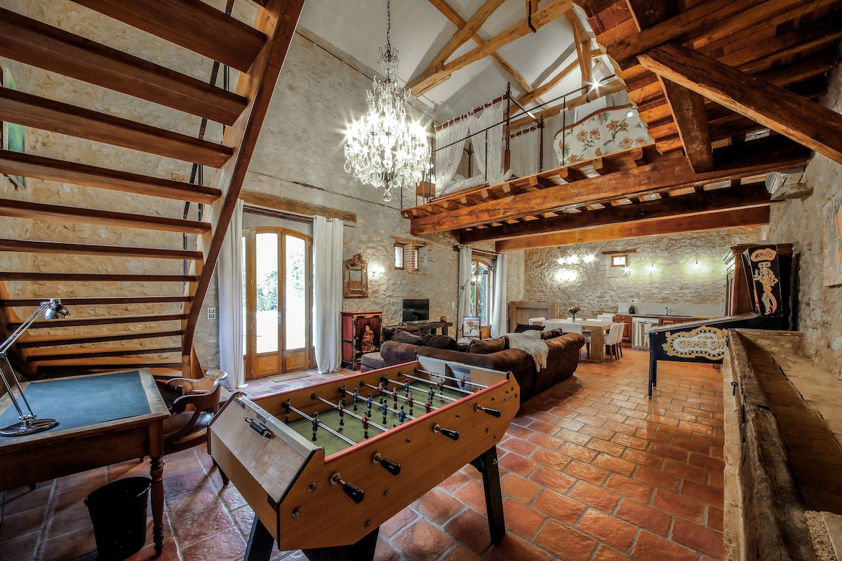 An open-plan living area features a pool table and foosball table, surrounded by comfortable seating. Exposed stone walls and wooden beams contribute to the rustic charm. Natural light enters through large doors, leading to a cozy dining space adorned with elegant lighting.