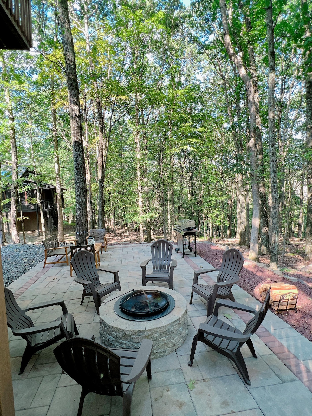 An outdoor setting is presented with a stone fire pit surrounded by several wooden chairs on a stone patio. Tall trees create a natural backdrop, providing a sense of privacy. A grill is visible nearby, offering additional outdoor cooking options.