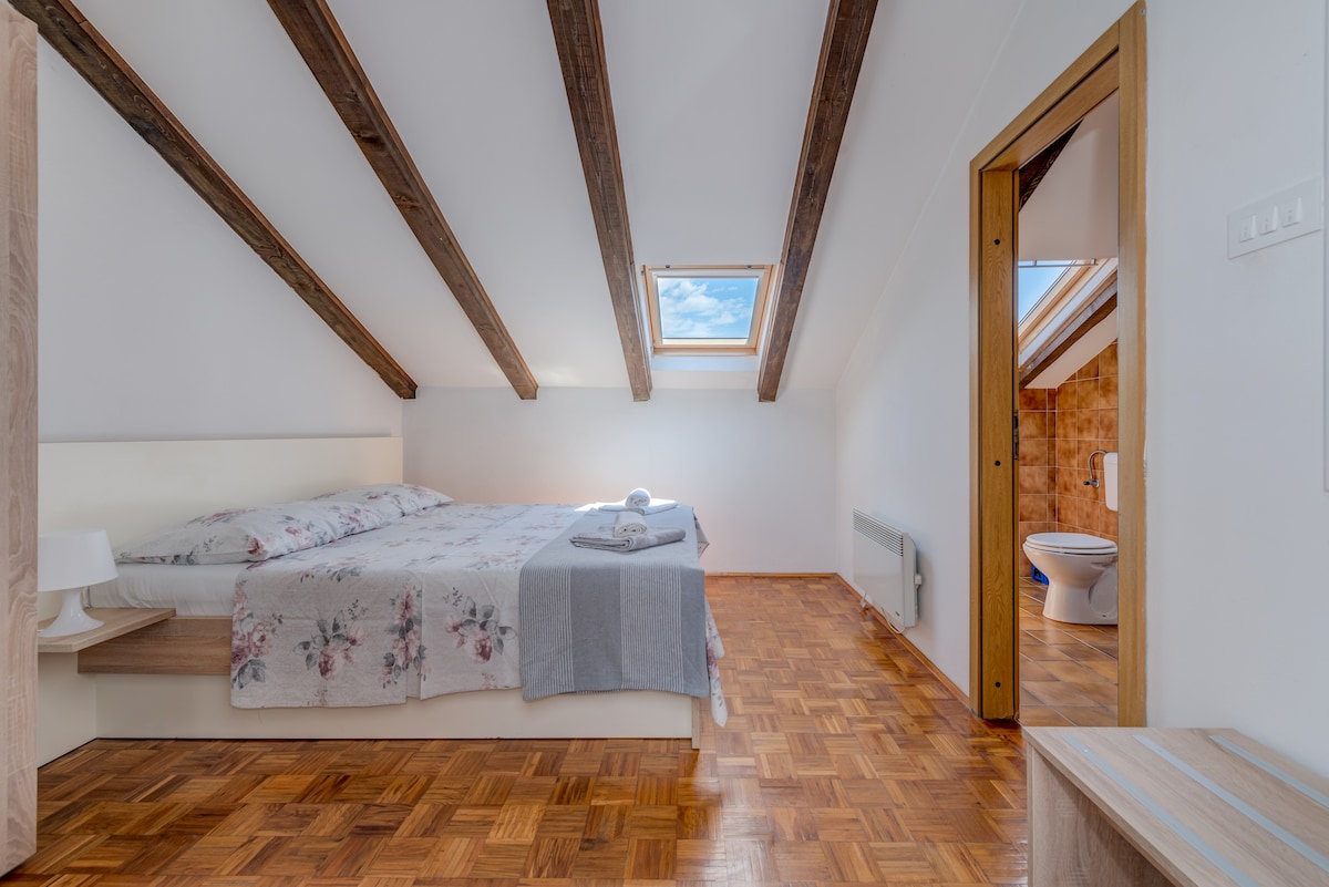 A cozy sleeping area is defined by sloped wooden beams overhead. A spacious bed with floral bedding is complemented by a soft blanket. Natural light enters through a skylight, illuminating the warm wooden floor. A bathroom door is visible in the background.