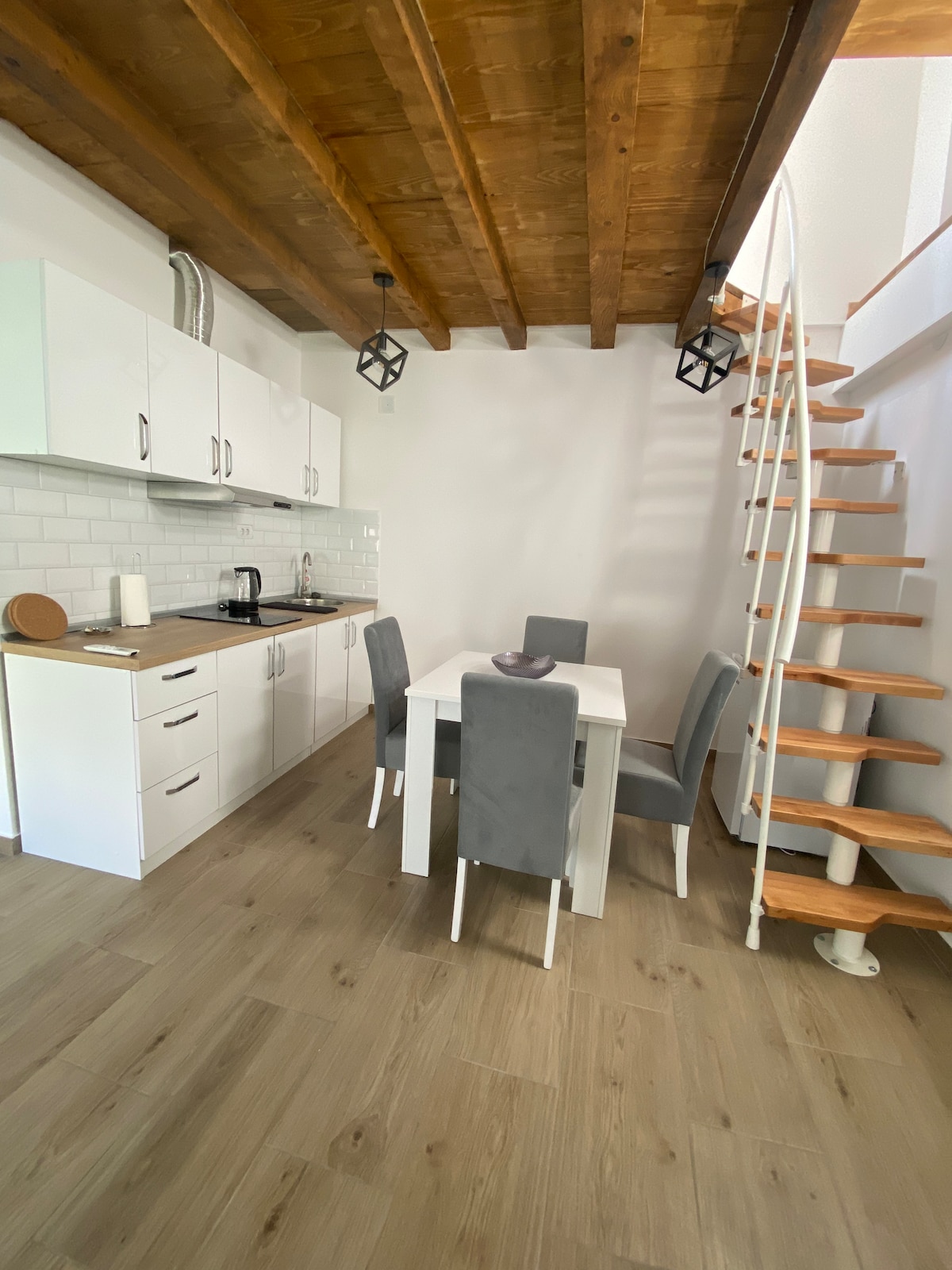 A contemporary kitchen area is presented with sleek white cabinetry, featuring a compact dining table surrounded by four chairs. A spiral staircase leads to the upper level. Bright light filters in, highlighting the wooden beams above and the warm tones of the flooring.