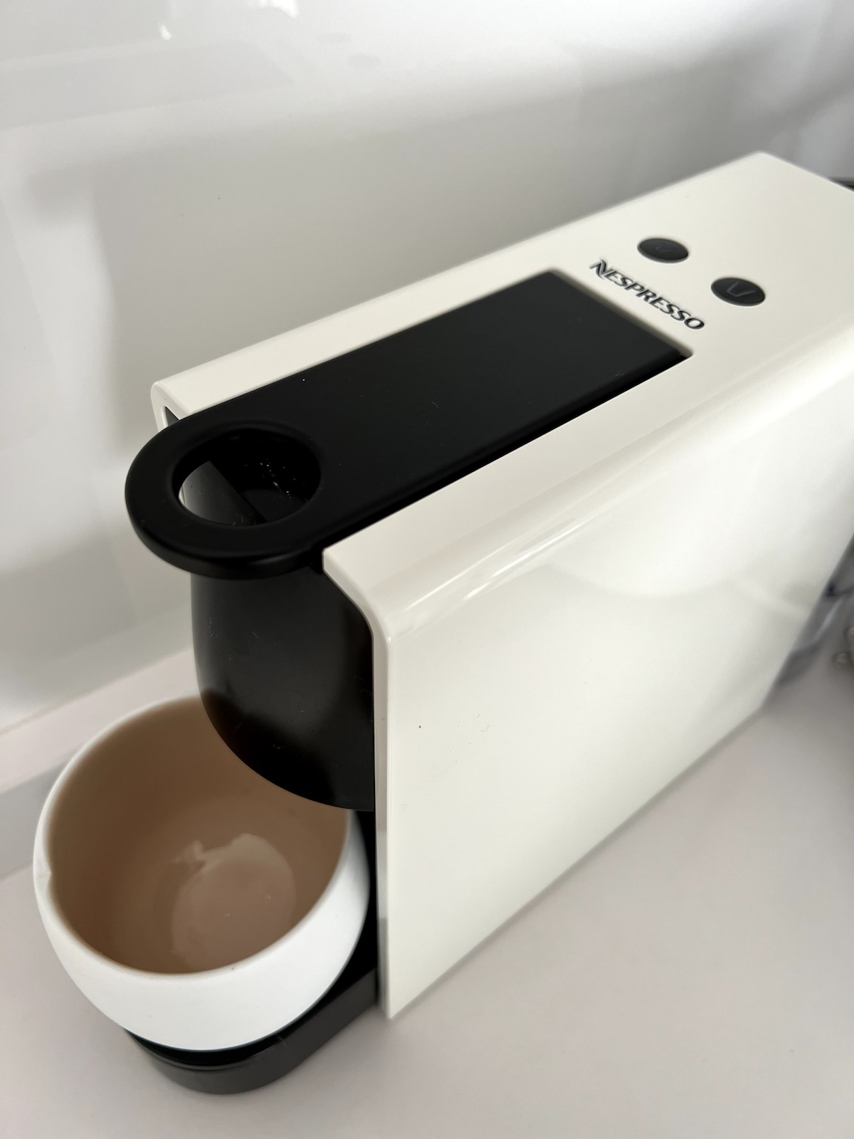 A Nespresso coffee machine is displayed in sleek white and black design, positioned on a smooth countertop. A small white cup sits beside it, ready for use. The machine features two buttons on its front for easy operation.
