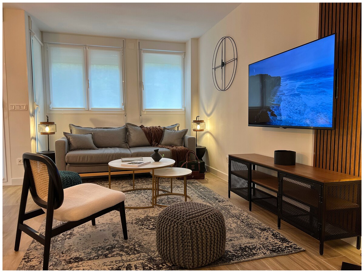 A modern living area features a comfortable gray sofa paired with a light-colored accent chair. A circular coffee table sits on a textured rug, while a large wall-mounted television displays a coastal scene. Natural light enters through large windows, brightening the space.