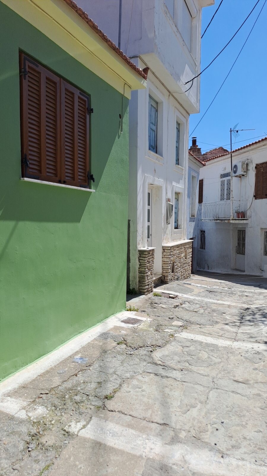 A narrow alleyway is bordered by two buildings, one featuring vibrant green walls and wooden shutters. Sunlight illuminates the stone path, highlighting the textures of the surrounding architecture. The area appears quiet and charming, with hints of nearby structures visible in the distance.