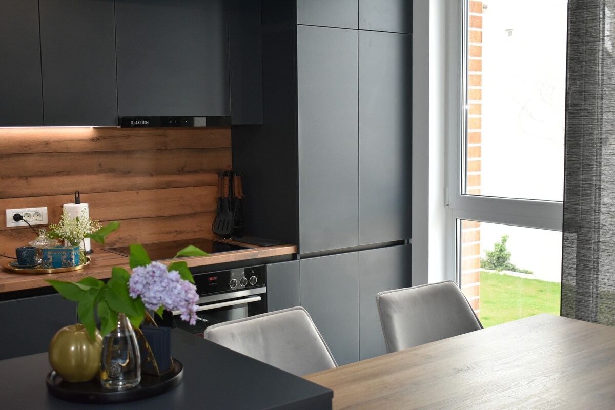 A modern kitchen is equipped with sleek black cabinetry and warm wooden accents. A dining table is set nearby, complemented by stylish chairs and decorative items. Natural light filters through a large window, illuminating the space and enhancing the greenery visible outside.
