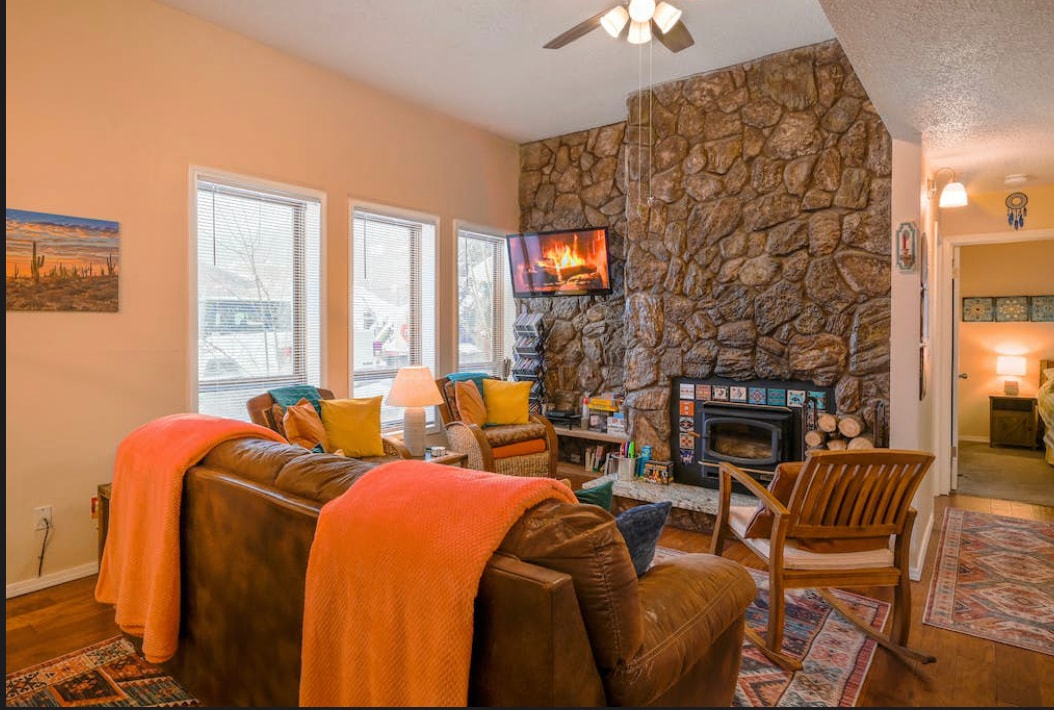A cozy living area is furnished with a leather sofa and a wooden armchair. A stone fireplace serves as a focal point, flanked by large windows allowing natural light. Colorful throw pillows and blankets add warmth to the space, complemented by an inviting artwork on the wall.