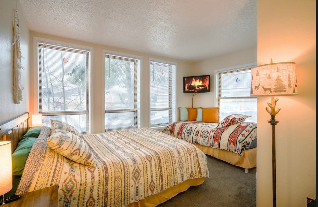 A bright bedroom features two spacious beds, each adorned with colorful, patterned bedding. Three large windows allow natural light to fill the room, with blinds partially drawn. A small wall-mounted fireplace provides warmth, and a floor lamp with a decorative shade enhances the cozy environment.