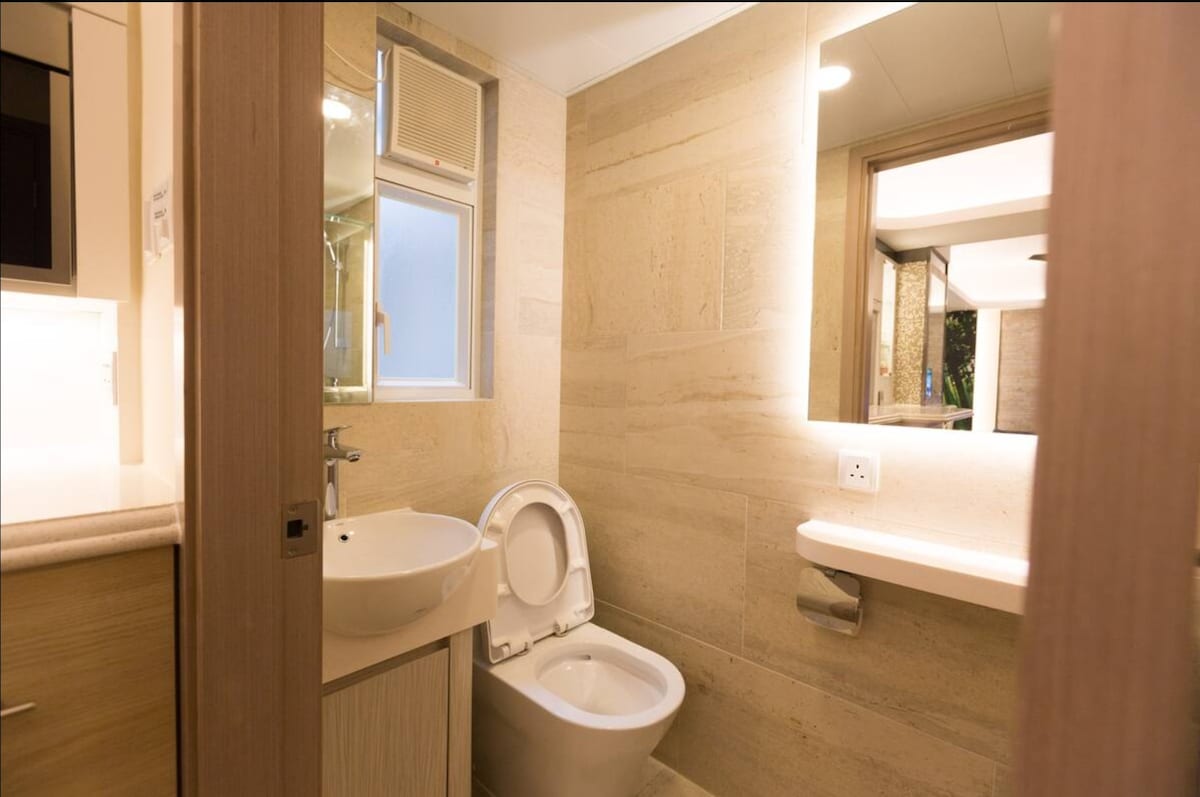 The bathroom features a clean design with beige stone walls and a modern sink adjacent to a toilet. A large mirror reflects the softly lit space, while a window provides natural light. A simple shelf holds toiletries, creating an organized and functional area.