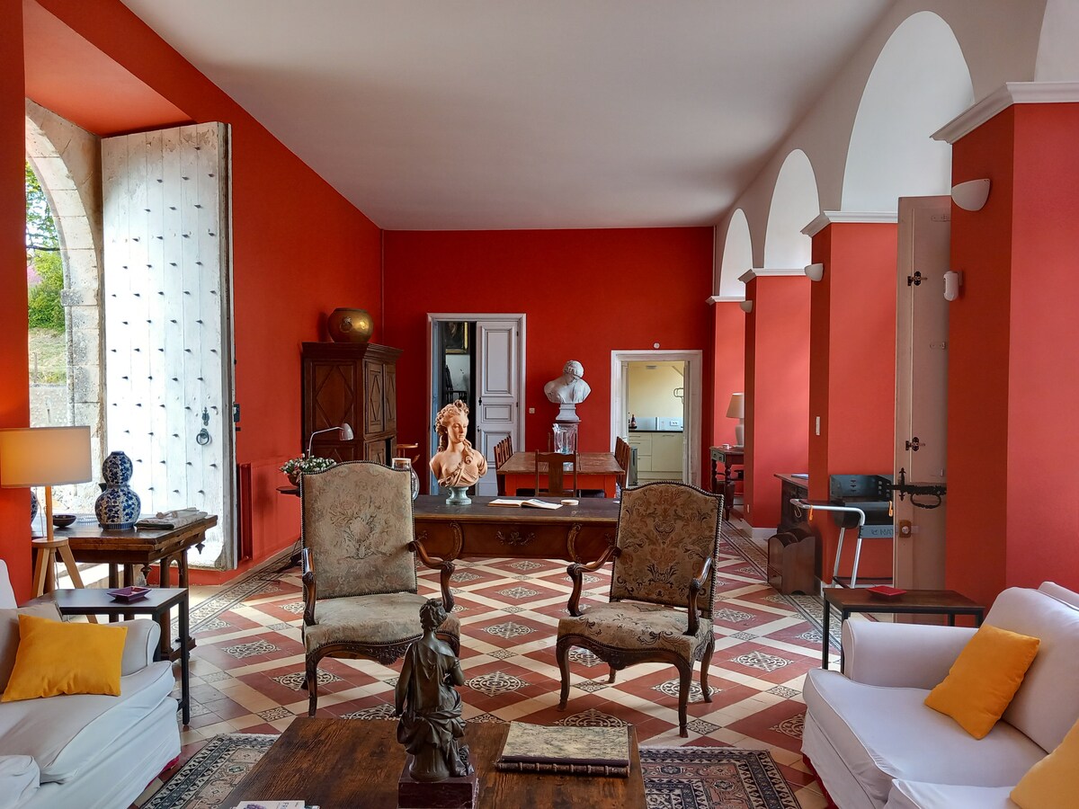 The spacious and bright living area features a rich red wall adorned with elegant arches. Antique furniture includes upholstered chairs and a wooden desk, complemented by decorative items. Large doors open to the outside, enhancing the room's airy feel and providing a view of the grounds.