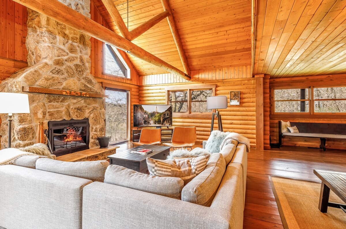 A spacious living area features a stone fireplace and large windows that provide natural light and views of the wooded surroundings. Comfortable seating is arranged around a coffee table, with warm wood finishes enhancing the inviting atmosphere. Decorative elements complement the rustic design.