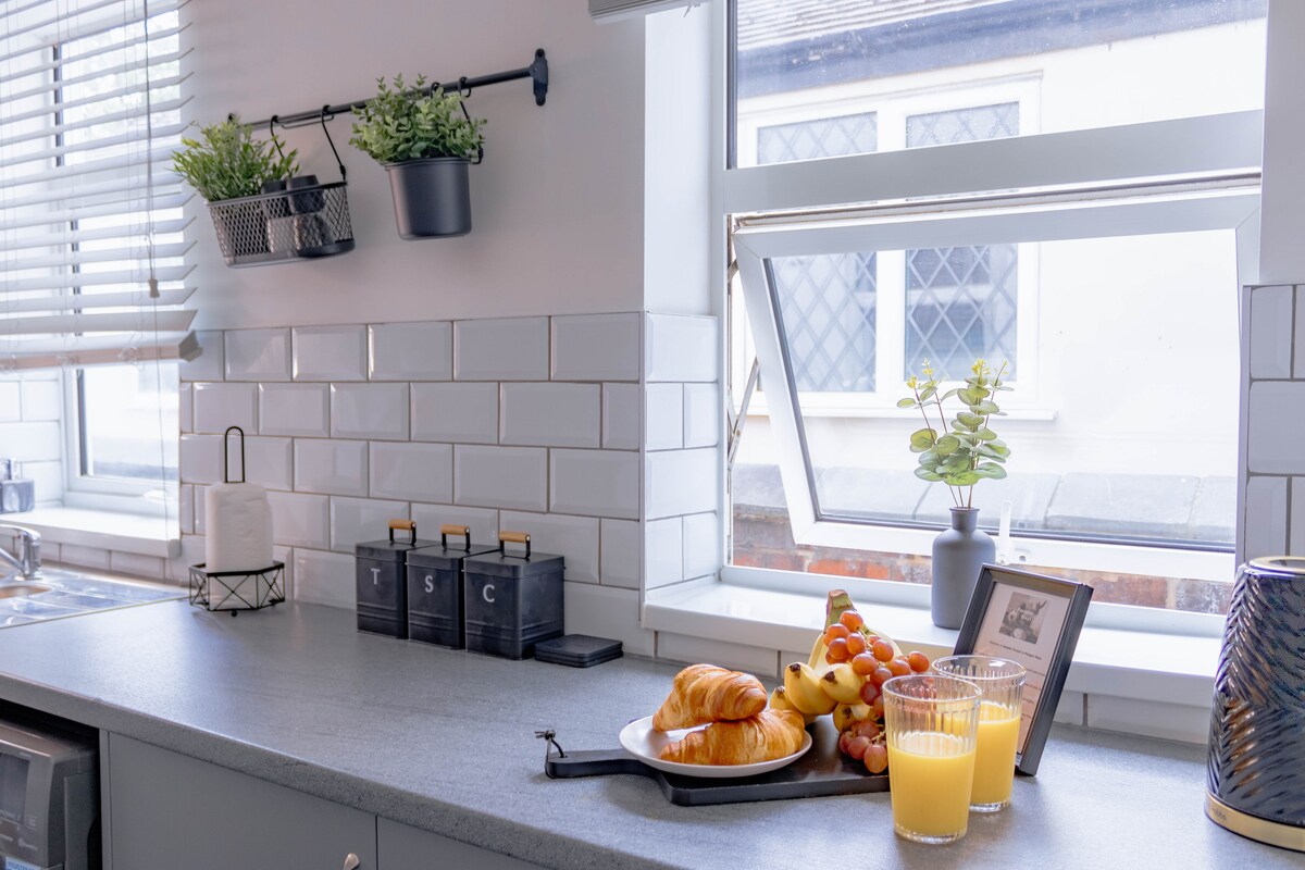 The kitchen countertop features a plate with a croissant and an assortment of fruits. Two glasses of orange juice sit beside a decorative photo frame. A small potted plant adds a touch of greenery, while hanging planters display additional plants. Natural light floods in through a partially opened window.