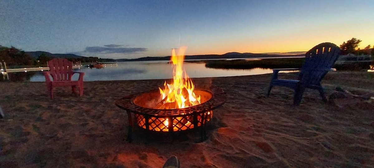A fire pit surrounded by colorful chairs is situated on the beach, with flames dancing in the center. The still lake reflects the twilight sky, while distant mountains provide a serene backdrop. Soft sand underfoot offers a comfortable seating area for evening gatherings.