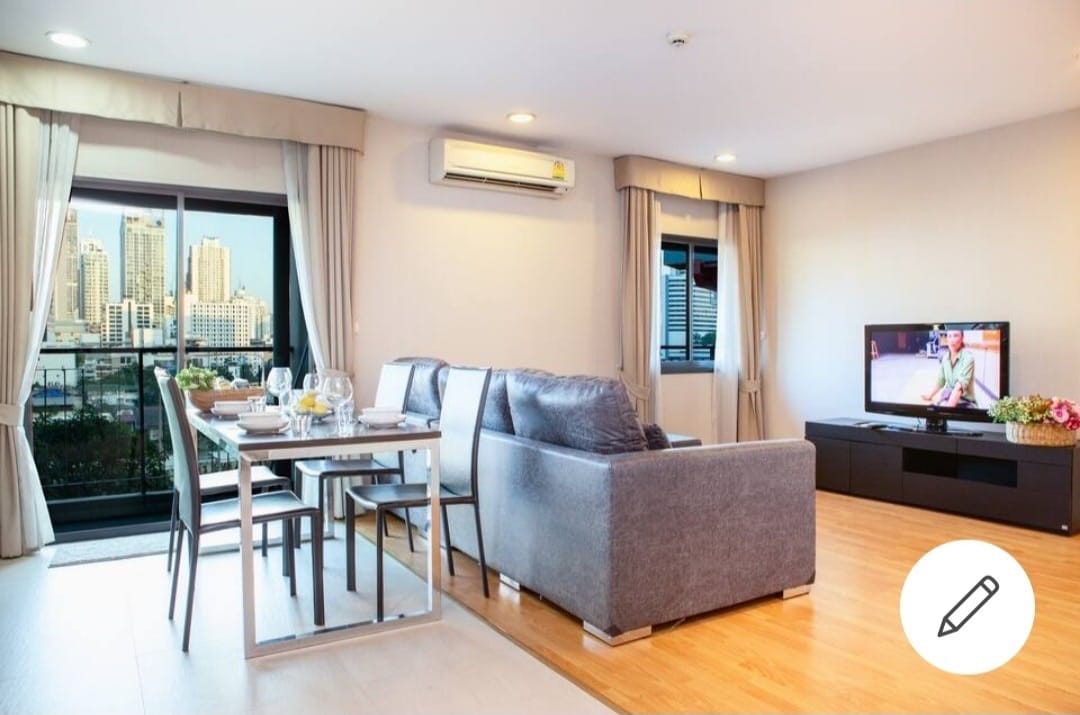 A modern living area is presented with a comfortable gray couch facing a television. A dining table set for four is positioned nearby, with a cityscape visible through large windows. Natural light brightens the space, complemented by light-colored curtains.