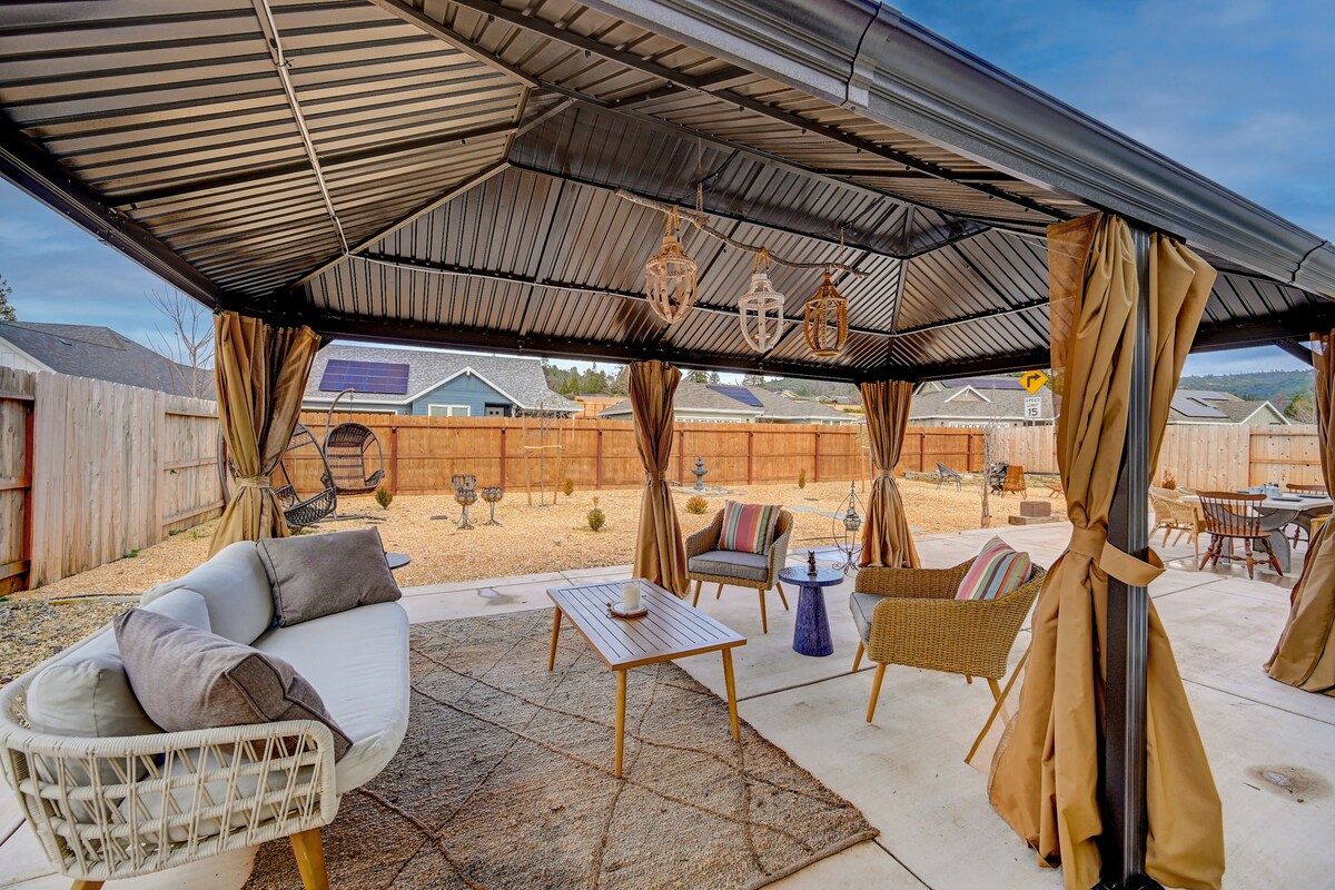 An outdoor living area is shaded by a large pergola, featuring comfortable seating arrangements on a concrete patio. Soft curtains frame the space, while a central coffee table adds functionality. The backyard is fenced, providing privacy and a serene atmosphere, with gravel underfoot.