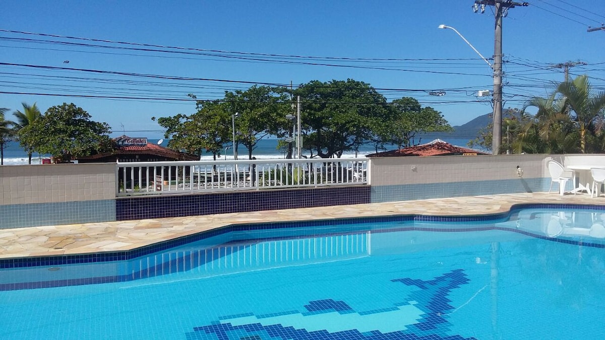The outdoor pool area features a clear blue pool surrounded by a stone deck. Lush palm trees and a distant beach are visible, providing a serene backdrop. A white railing outlines one side, with several white chairs positioned nearby.