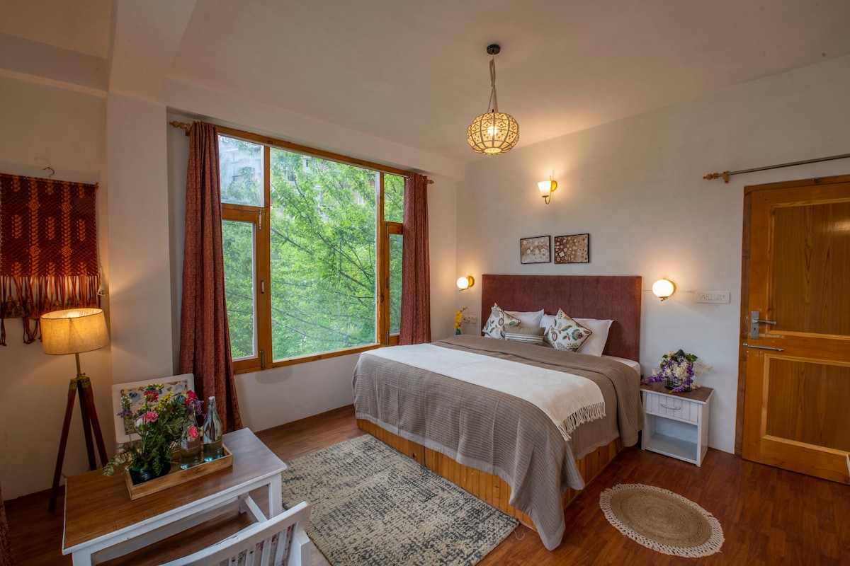 A spacious bedroom is illuminated by natural light filtering through large windows. A comfortable bed is positioned against a wall, complemented by decorative pillows. A small wooden table with floral accents and a soft rug adds warmth to the wooden flooring.