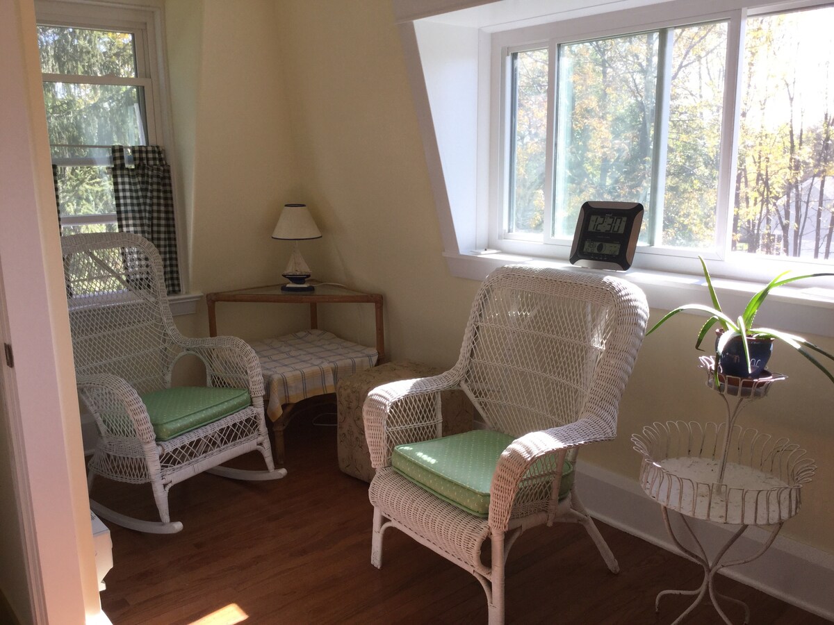 A sunlit corner features two white wicker rocking chairs with green cushions, positioned beside a small table. A lamp sits on the table, and a decorative plant is displayed in a white planter. Large windows allow natural light to fill the space.