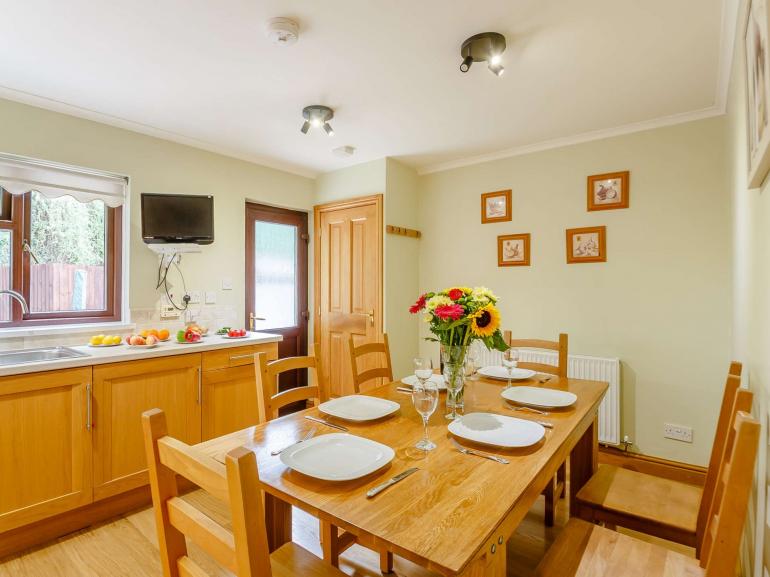 A light-filled dining area features a wooden table set for six, complete with white plates and glassware. Sunflowers and other flowers are arranged attractively in a vase. Storage cabinets and a television are visible, alongside natural wood accents throughout the space.