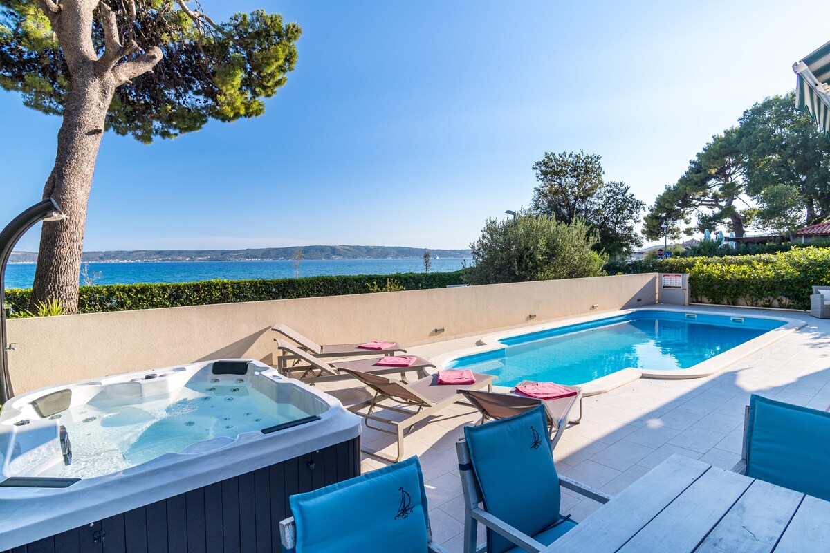 An outdoor space features a private swimming pool and a jacuzzi, surrounded by sun loungers and a scenic view of the sea. Lush greenery frames the area, while blue skies enhance the bright atmosphere. Sunbeds are positioned for relaxation by the poolside.