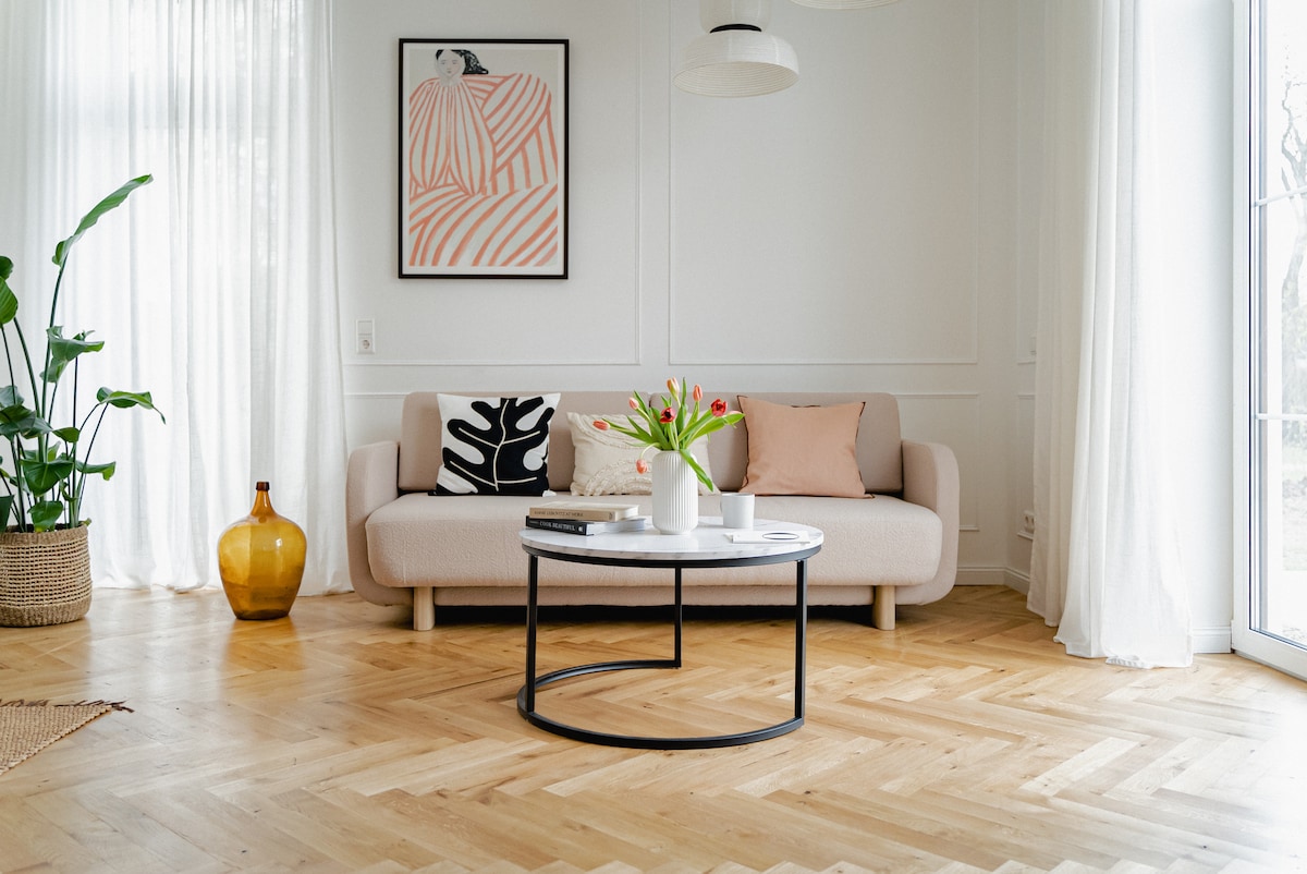 A minimalist living area features a light-colored sofa adorned with decorative pillows. A round coffee table, set centrally, holds a vase with fresh flowers. Natural light floods the room through large windows, highlighting the warm wooden herringbone floor and greenery in the background.