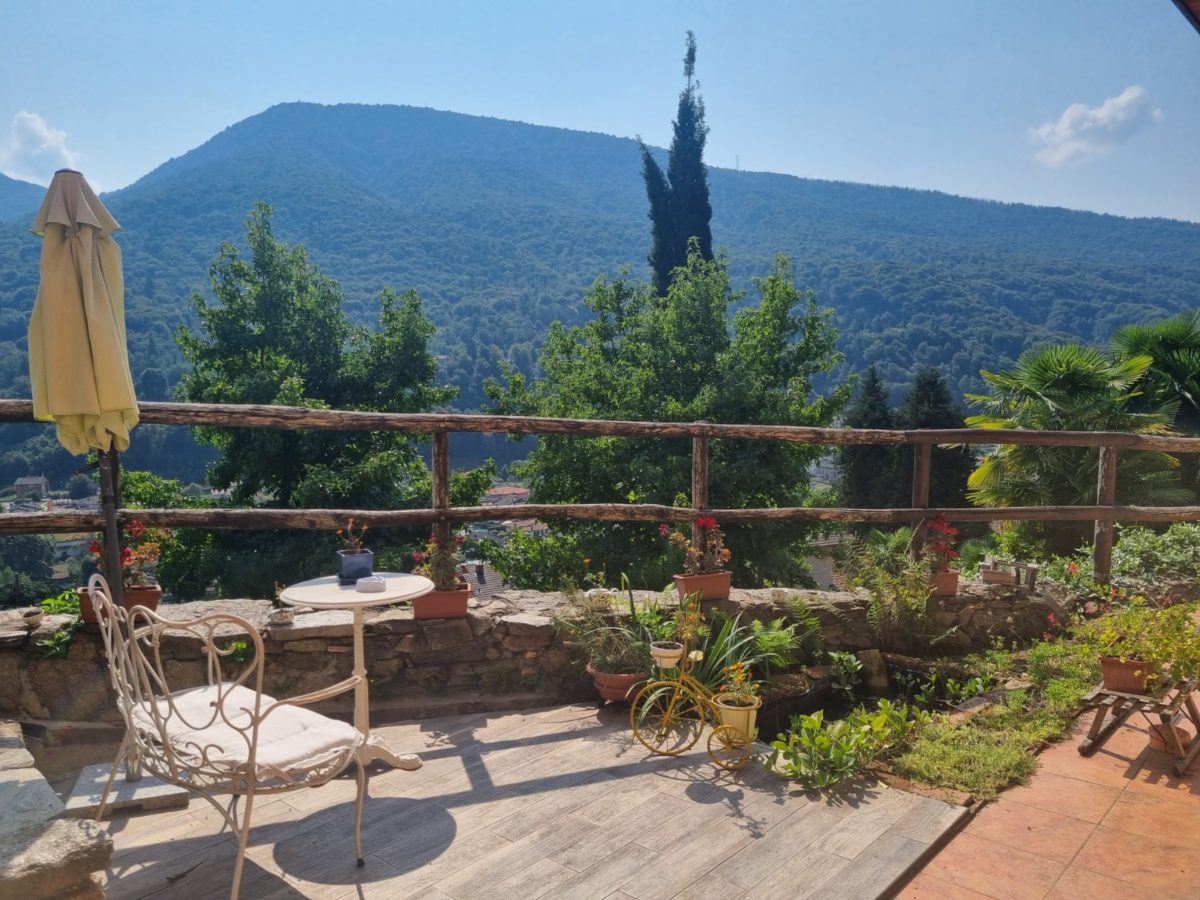 A scenic patio area features a small round table with two chairs, surrounded by potted plants. A large umbrella provides shade. In the background, a lush green mountain is visible under a clear blue sky, enhancing the peaceful outdoor setting.